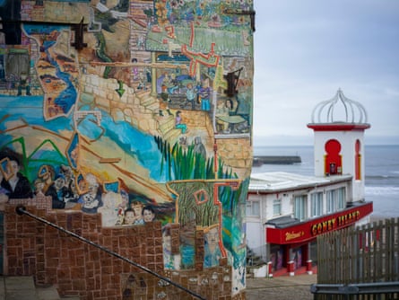 A mural featuring children’s and other faces, and scenes from the town on a wall alongside steps leading to an old-fashioned arcade on the seafront