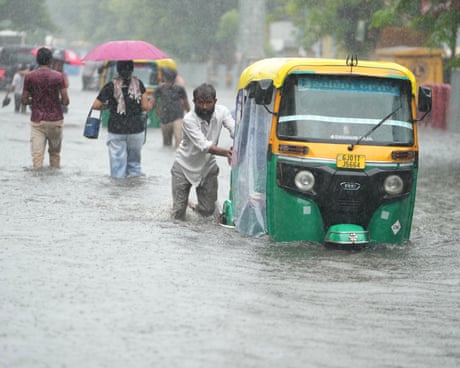 People wade through a flooded street and a man pushes a stalled motor rickshaw