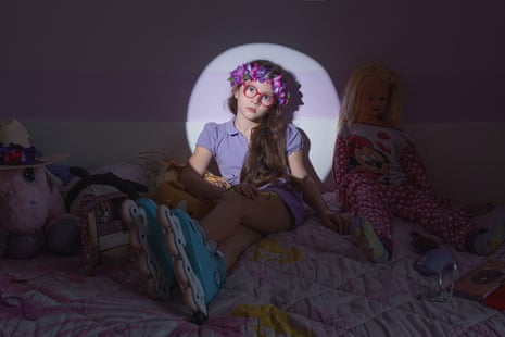 Giulia is spotlit in a light circle in her dark bedroom to focus on her head and shoulders; she wears red-framed glasses and a floral wreath around her head, plus a lilac-coloured short-sleeved top.