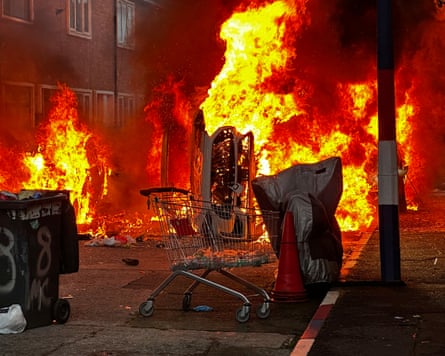 A car on fire in front of a larger fire in a residential street.