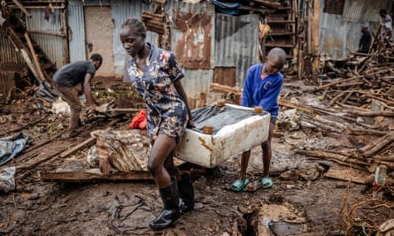 A girl and a boy carry a piece of furniture across muddy, debris-strewn ground