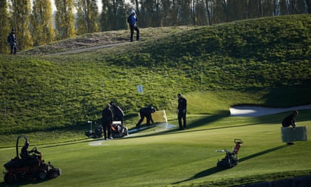 Greenkeepers work before the Ryder Cup at Le Golf National Course at Saint-Quentin-en-Yvelines, south-west of Paris: mowers, sprinklers and other equipment can be seen on a perfectly cropped green beneath a grassy bank with tall trees in the background