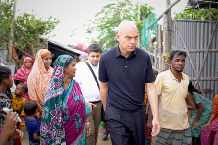 A bald European man walks past corrugated-iron shelters with a Bangladeshi woman in a sari and a man in a lungi