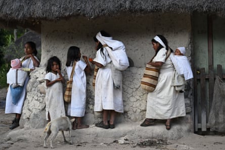 People dressed in white stand around a building that appears to have mud walls and a thatched roof