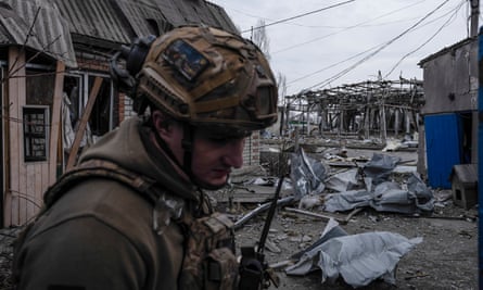 A soldier in helmet and fatigues stands in front of ruined buildings, looking downward