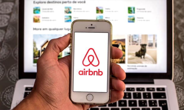 The Airbnb app seen displayed on a smartphone screen with the Airbnb website displayed on a laptop in the background. 
