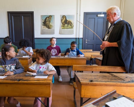 Children sitting at wooden desks watched over by a teacher in a black gown and holding a cane