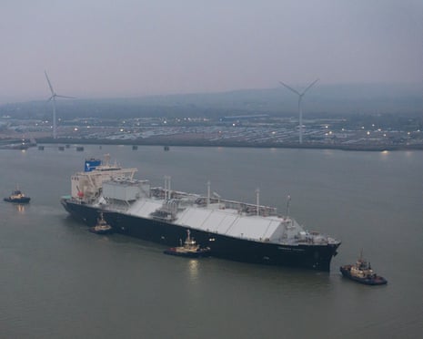 An LNG tanker arrives at the Isle of Grain terminal, escorted by tugboats, wind turbines beyond