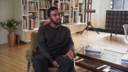 Penuel Mlotshwa sit sin an airy modern room next to a coffee table and in front of a full bookshelf.