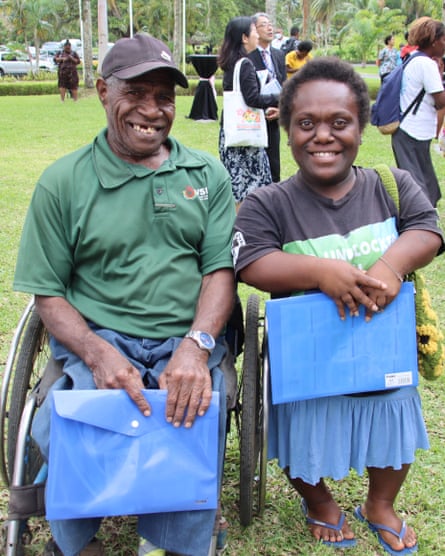 Arthur Simrai sits in his wheelchair smiling broadly. Standing next to him is a person of shorter stature
