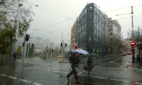 A view of people crossing a city street through a rain covered window. The pedestrians are carrying umbrellas and there are powerlines over head in front of a multi-storey building