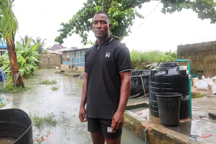 An African man in a black polo shirt and shorts standing among various tanks and outbuildings with flood water visible in the yard.