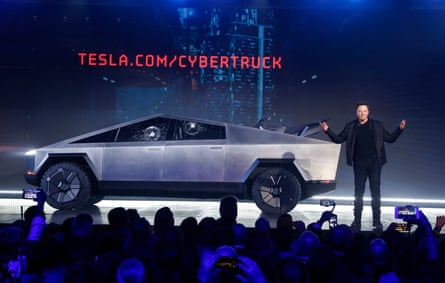 A man wearing all-black speaks on stage next to a futuristic-looking truck with cracks in the window as a crowd looks on and takes photos