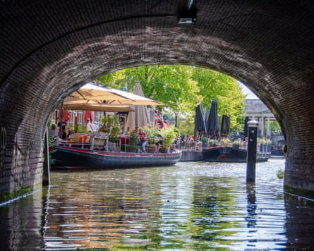 View through the arched brick bridge over a canal on to houseboats and, behind them, parasols, tables and chairs