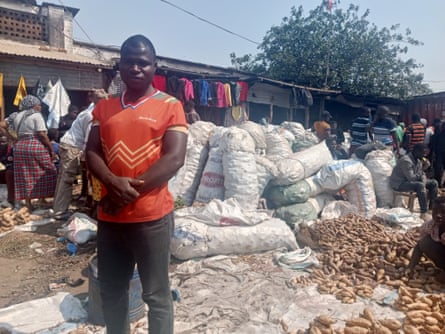 A man in a red T-shirt stands in front of piled-up bags and sweet potatoes spread out on the ground