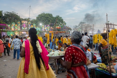 Festival goers walk past fruit stalls towards a display of neon light decorations at dusk