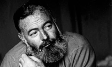 Ernest Hemingway, in a slightly wrinkled shirt and wearing a leather-strapped watch – his beard dark and long and with grey streaks – holds his hand near his face and smiles slightly