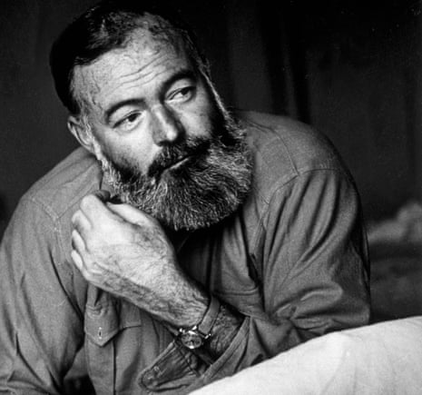 Ernest Hemingway, in a slightly wrinkled shirt and wearing a leather-strapped watch – his beard dark and long and with grey streaks – holds his hand near his face and smiles slightly