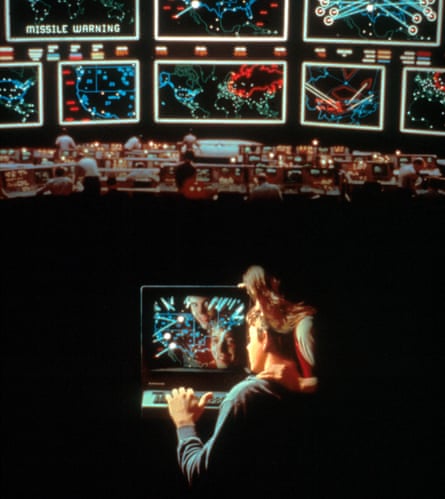 Two teenagers look at a computer screen with a huge video of bank of missile warning screens above them.
