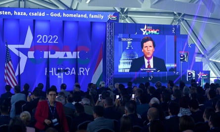 A crowd watches a giant screen where Tucker Carlson can be seen delivering a speech.