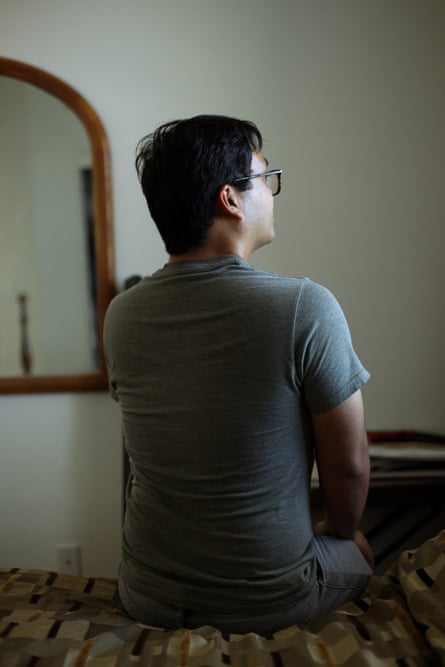 the back of a young man wearing glasses and a T-shirt sitting on the edge of a bed