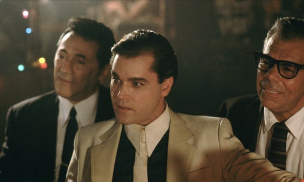 Liotta in a white suit, black shirt and white tie talking to someone off camera next to two hemlines in black suits