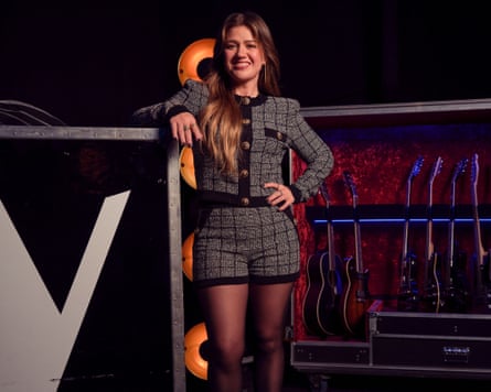 Kelly Clarkson in a tweed outfit smiles, leaning near a row of guitars onstage