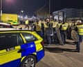 Officers in yellow hi-vis vests holding shields and batons stand by a police car with a crowd in the background. It is dark and they are in front of port buildings.