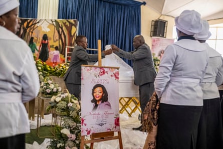 Women in white tunics and hats stand near a coffin as men remove a lace covering from it