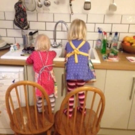 Two girls, backs to the camera, stand on chairs to wash up at the kitchen sink