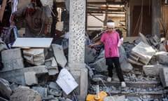 A man lifts rubble while a child with a bandaged head walks down rubble-strewn steps in a bombed house