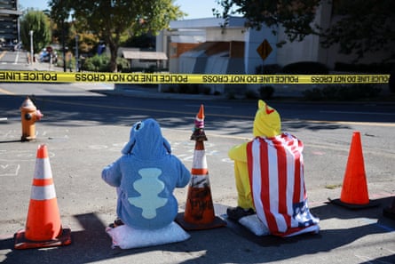 Two people, one dressed as Stitch and the other as a chicken (draped in a US flag) sit on white pillows between orange cones and beneath yellow tape.