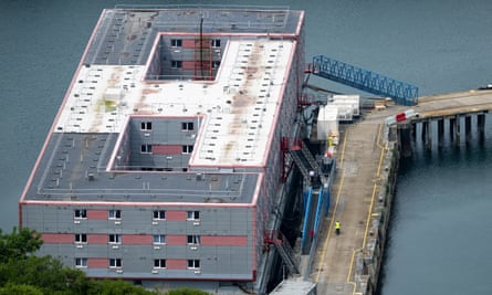 overhead view of the Bibby Stockholm barge, moored in Portland harbour: it is a series of square grey accommodation blocks with tiny windows, mounted on pontoons, with access via footbridges from the dock.