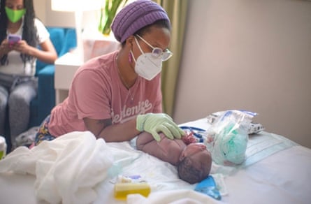 A woman in a pink T-shirt, purple hat, glasses, and surgical mask, holds her gloved hands over the folded arms and legs of a newborn baby on a bed.
