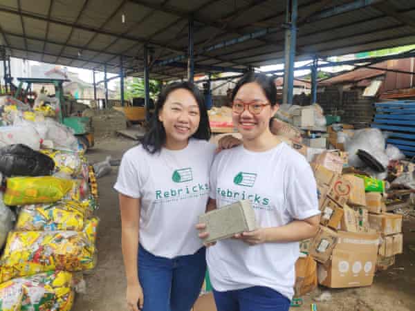 Ovy Sabrina and Novita Tan stand in a warehouse full of waste plastic. Tan is holding a brick made of recycled plastic and cement.