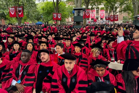 A large crowd of graduates wearing red and black gowns and mortarboards gathers outside Harvard University