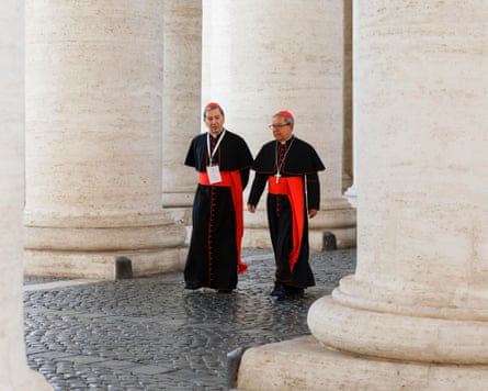 Two cardinals in black robes with red hats and red belts walk past stone columns in Vatican City