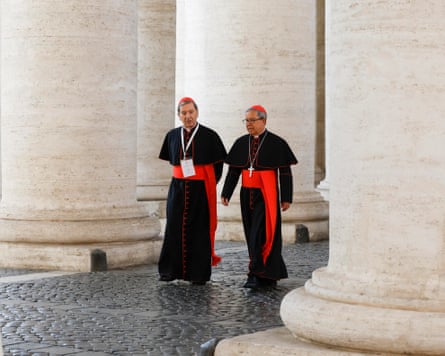 Two cardinals in black robes with red hats and red belts walk past stone columns in Vatican City
