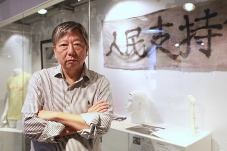 Lee Cheuk-Yan stands in front of a display with a banner and figurine with his arms folded; he appears to be in his 60s with greying hair.