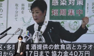 A man wearing a protective mask walks in front of a public tv screen showing Tokyo governor, Yuriko Koike, speaking at a press conference