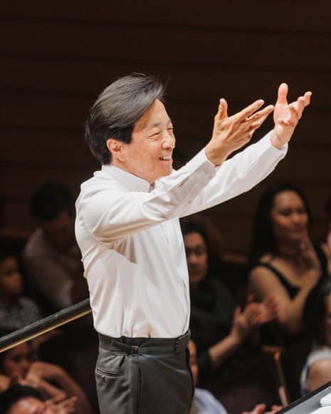 Cheung stands in front of an unseen orchestra with crowd applauding behind him as he indicates to the orchestra to stand for applause