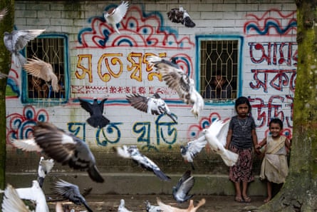 Pigeons fly in front of two children standing in front of a building with what appears to be Bengali script written on it