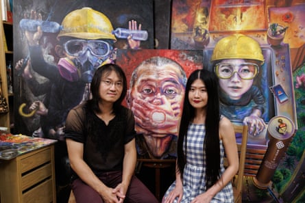 Lumli and Lumlong, political artists, sit in front of paintings in their studio that feature protesters with yellow hard hats and other scenes of protest and another with a man with two hands clamped over his mouth.