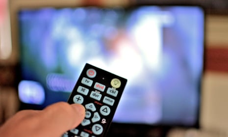 Closeup view of a television remote in hand with the tv blurred in the background.