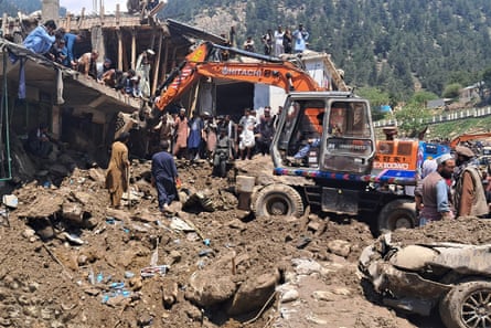 An excavator is used to clear away mud and debris from a damaged building, as people look on