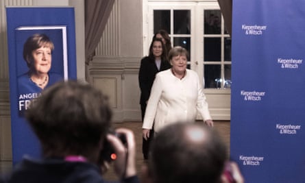 Merkel in white (centre) with woman standing behind, walks on to stage with image of her book cover (left) with several backs of heads in foreground