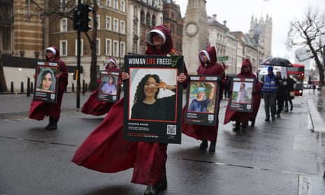 Women dressed as Handmaids march with placards that have images of female protesters detained or killed in Iran.