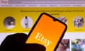 A hand holding a phone screen displaying the Etsy logo, in front of the Etsy website on a desktop screen