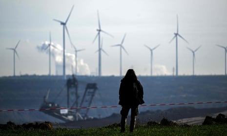 A woman facing aeway from the camera, silhouetted against the background, in front of a large pit with a minign machine in it, and a row of wind turbines on the horizon