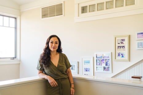 Woman stands on split level beyond what appears to be a stairwell, smiling, with sunny window behind her